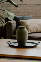 Green smooth memorial urn on a black tray with a couch and plant in the background