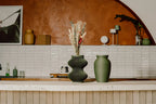 Decorative memorial urn beside a flower vase on a kitchen counter with a tiled wall and framed picture in the background