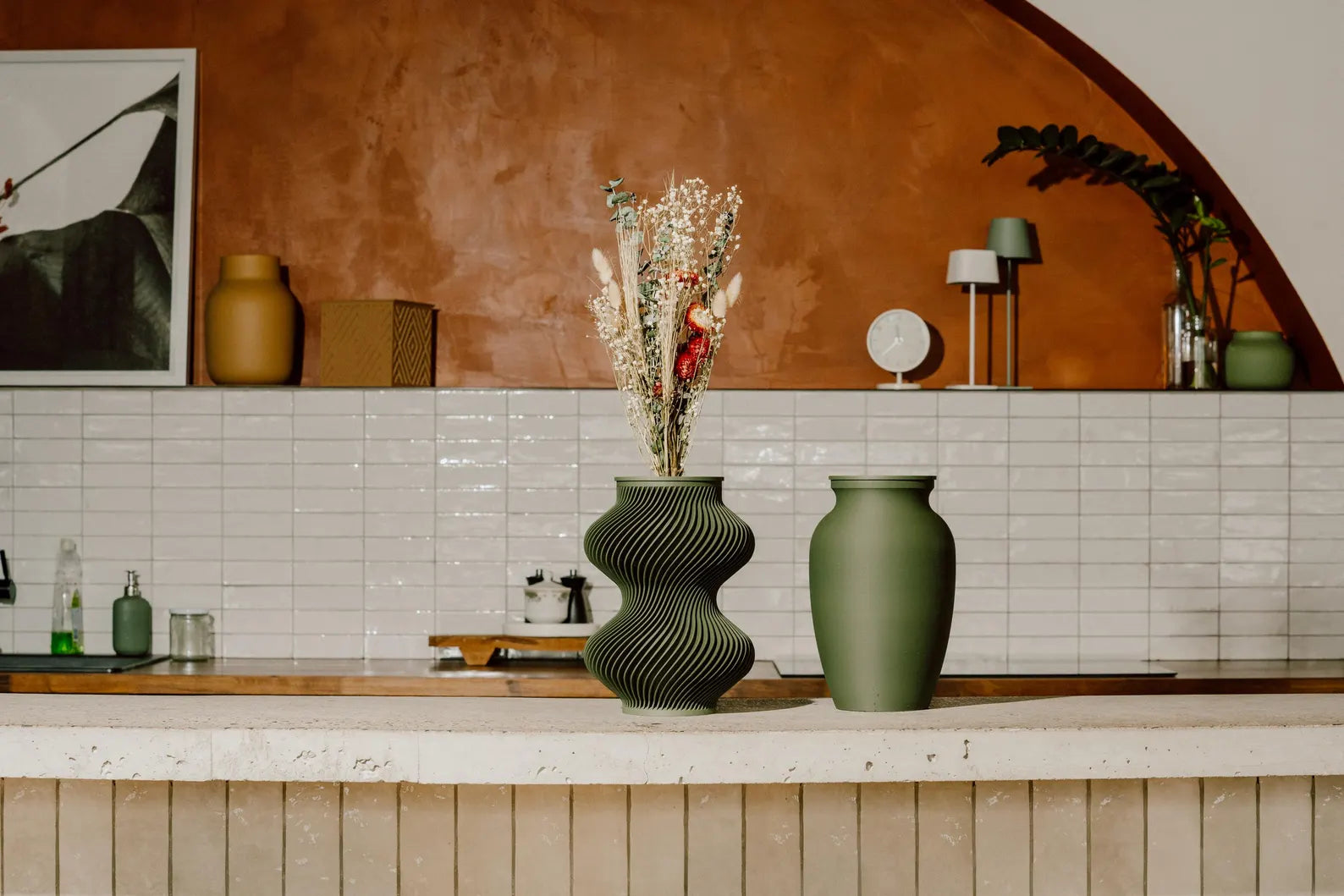 Decorative memorial urn beside a flower vase on a kitchen counter with a tiled wall and framed picture in the background