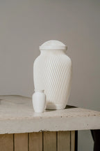 White ribbed memorial urn and smaller white urn on a wooden surface with a neutral background