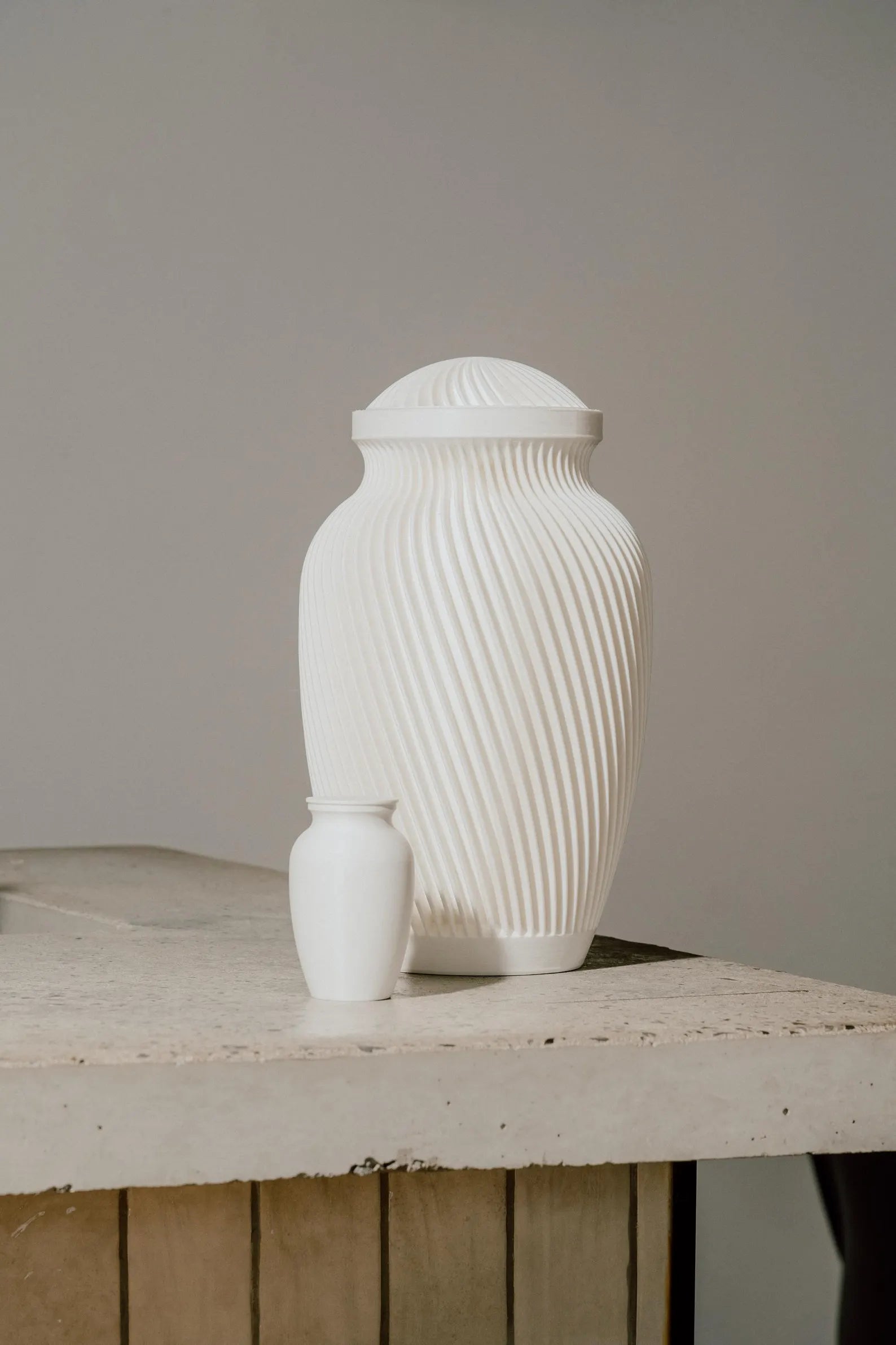 White ribbed memorial urn and smaller white urn on a wooden surface with a neutral background